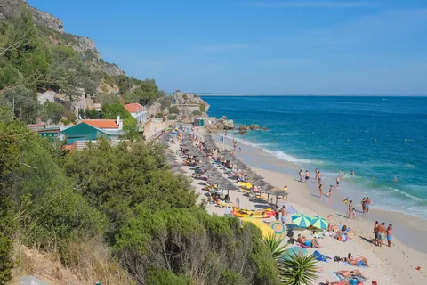 A bustling summer day at Portinho da Arrábida beach with sunbathers and umbrellas along the shore.